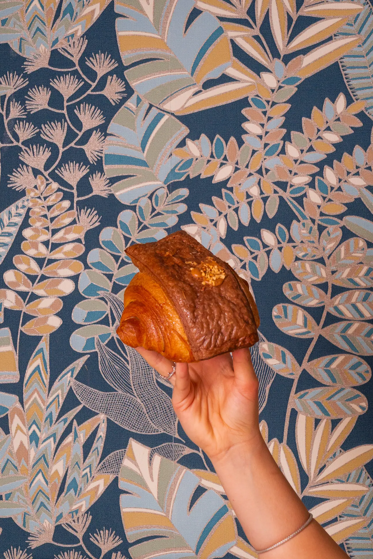 Una mano regge un croissant tondo davanti alla carta da parati a foglie